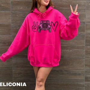 May include: A bright hot pink hooded sweatshirt featuring a graphic print on the chest. The design includes a black peace sign, a black outline of a heart, and a central illustration of a dark-colored off-road vehicle with a rear-mounted spare tire.