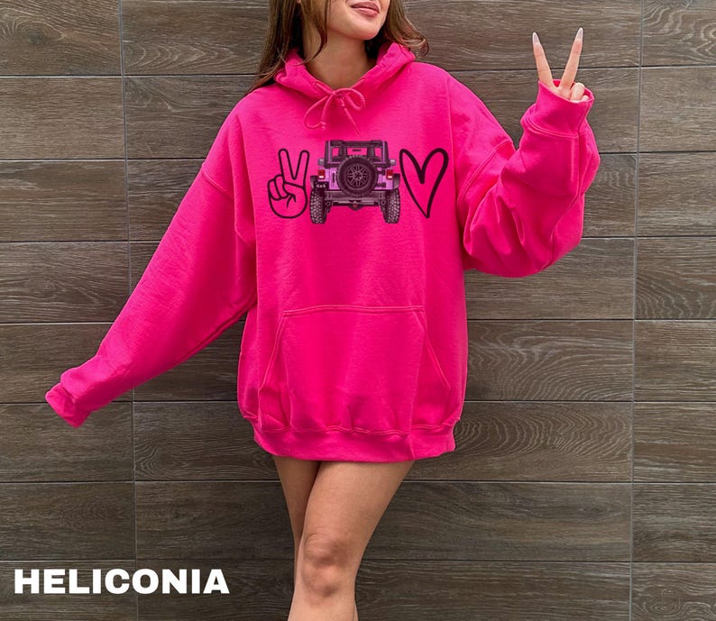 May include: A bright hot pink hooded sweatshirt featuring a graphic print on the chest. The design includes a black peace sign, a black outline of a heart, and a central illustration of a dark-colored off-road vehicle with a rear-mounted spare tire.
