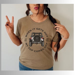 Peut inclure: Un t-shirt vert olive chin&eacute; avec une illustration de l'arri&egrave;re d'un v&eacute;hicule tout-terrain blanc avec une roue de secours. Le texte "WHEN IT GETS HOT MY TOP COMES OFF" est dispos&eacute; en cercle, accompagn&eacute; de deux ic&ocirc;nes de signe de la paix.