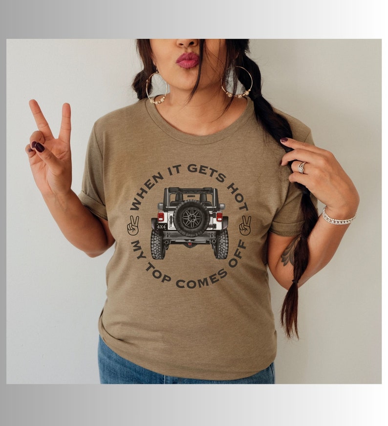 Peut inclure: Un t-shirt vert olive chin&eacute; avec une illustration de l'arri&egrave;re d'un v&eacute;hicule tout-terrain blanc avec une roue de secours. Le texte "WHEN IT GETS HOT MY TOP COMES OFF" est dispos&eacute; en cercle, accompagn&eacute; de deux ic&ocirc;nes de signe de la paix.