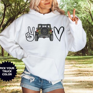May include: A white hooded sweatshirt featuring a graphic print on the front. The design includes a black peace sign, a detailed illustration of an olive green off-road vehicle viewed from the rear, and a black heart. A black starburst icon says PICK YOUR TRUCK COLOR.