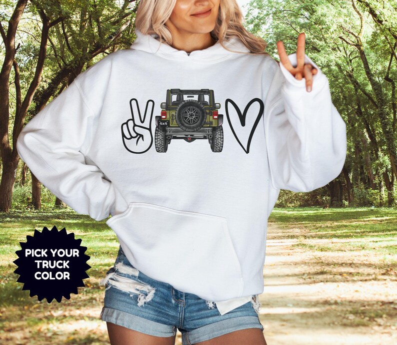 May include: A white hooded sweatshirt featuring a graphic print on the front. The design includes a black peace sign, a detailed illustration of an olive green off-road vehicle viewed from the rear, and a black heart. A black starburst icon says PICK YOUR TRUCK COLOR.