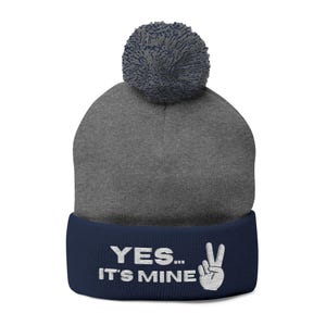May include: A charcoal gray knit beanie with a dark blue pom-pom on top. The beanie features a wide dark blue cuff with the white embroidered text "YES... IT'S MINE" and a white embroidered hand gesture showing a peace sign on the front.