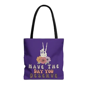 May include: Purple tote bag with a skeleton hand giving the peace sign with flowers around it. The text on the bag reads "Have the day you deserve."
