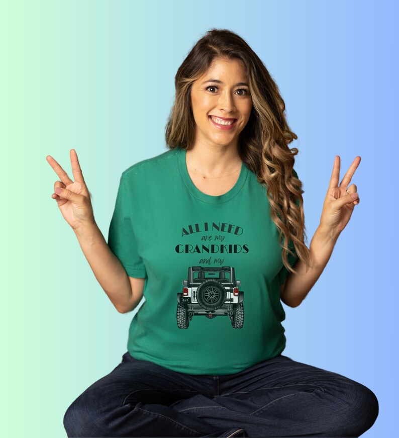 Puede incluir: Una mujer con una camiseta verde con el texto "All I need are my grandkids and my" y una ilustraci&oacute;n en blanco y negro de un Jeep. Est&aacute; sonriendo y haciendo el signo de la paz con ambas manos.