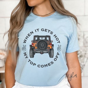 Peut inclure: Un t-shirt bleu clair &agrave; manches courtes avec un motif repr&eacute;sentant un v&eacute;hicule tout-terrain orange vu de l'arri&egrave;re. Le texte "WHEN IT GETS HOT MY TOP COMES OFF" est imprim&eacute; en cercle autour du v&eacute;hicule, encadr&eacute; par deux signes de paix.