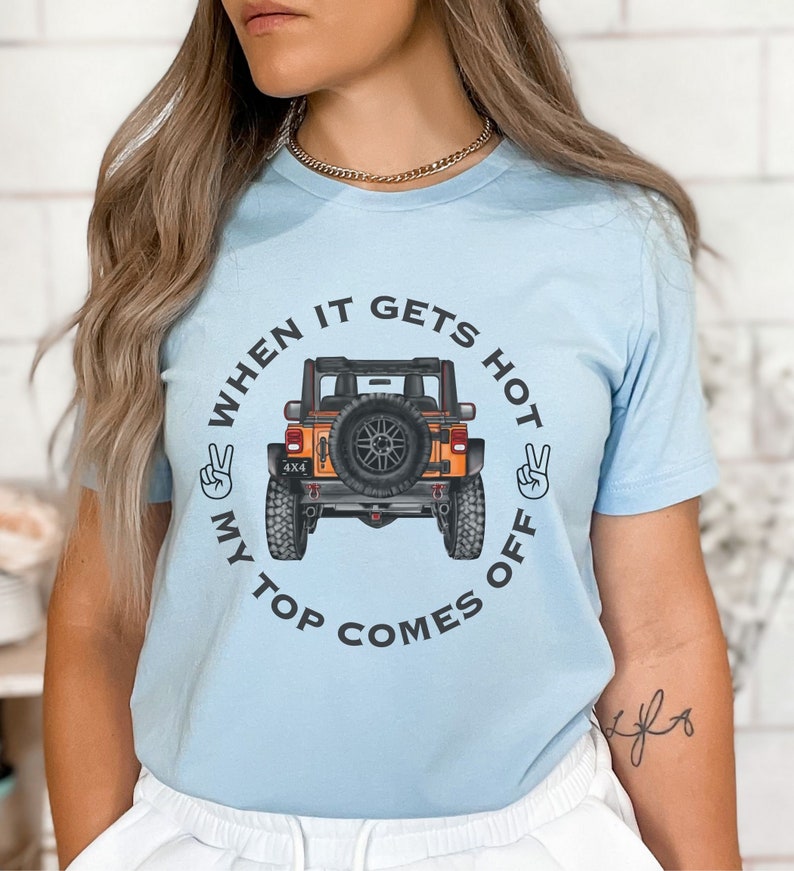 Peut inclure: Un t-shirt bleu clair &agrave; manches courtes avec un motif repr&eacute;sentant un v&eacute;hicule tout-terrain orange vu de l'arri&egrave;re. Le texte "WHEN IT GETS HOT MY TOP COMES OFF" est imprim&eacute; en cercle autour du v&eacute;hicule, encadr&eacute; par deux signes de paix.