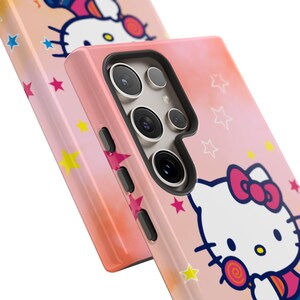 May include: Two pink phone cases with a Hello Kitty design. The cases have a gradient background with stars and a cartoon image of Hello Kitty. The phone cases have a black camera module and side buttons.