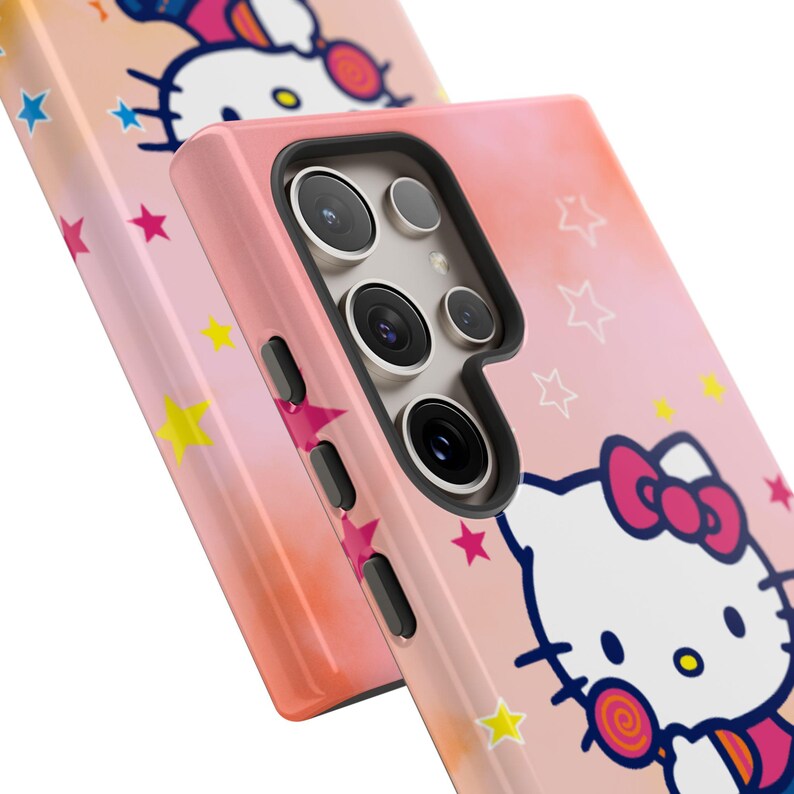 May include: Two pink phone cases with a Hello Kitty design. The cases have a gradient background with stars and a cartoon image of Hello Kitty. The phone cases have a black camera module and side buttons.