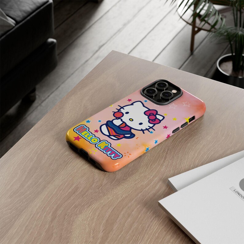 May include: A phone case with a Hello Kitty design. The case has a pink and orange gradient background with stars and the words "Hello Kitty". The design includes a cartoon image of Hello Kitty wearing a blue outfit and a pink bow.