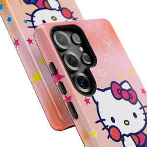 May include: Pink phone case featuring a Hello Kitty design with a bow and lollipop. The case has a gradient colour scheme and is decorated with stars. The phone case has a camera and side buttons.