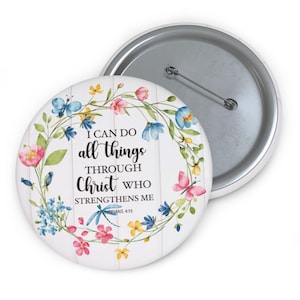 May include: A round white button with a floral wreath design featuring pink, blue, and yellow flowers, butterflies, and the text "I CAN DO all things THROUGH CHRIST who STRENGTHENS ME" from Philippians 4:13. The button has a metal pin back.
