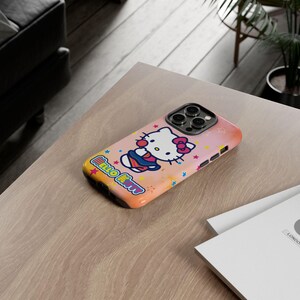 May include: A pink and orange iPhone case featuring Hello Kitty. The case has a colourful design with stars and the words "Hello Kitty". The phone case is on a light brown wooden table.