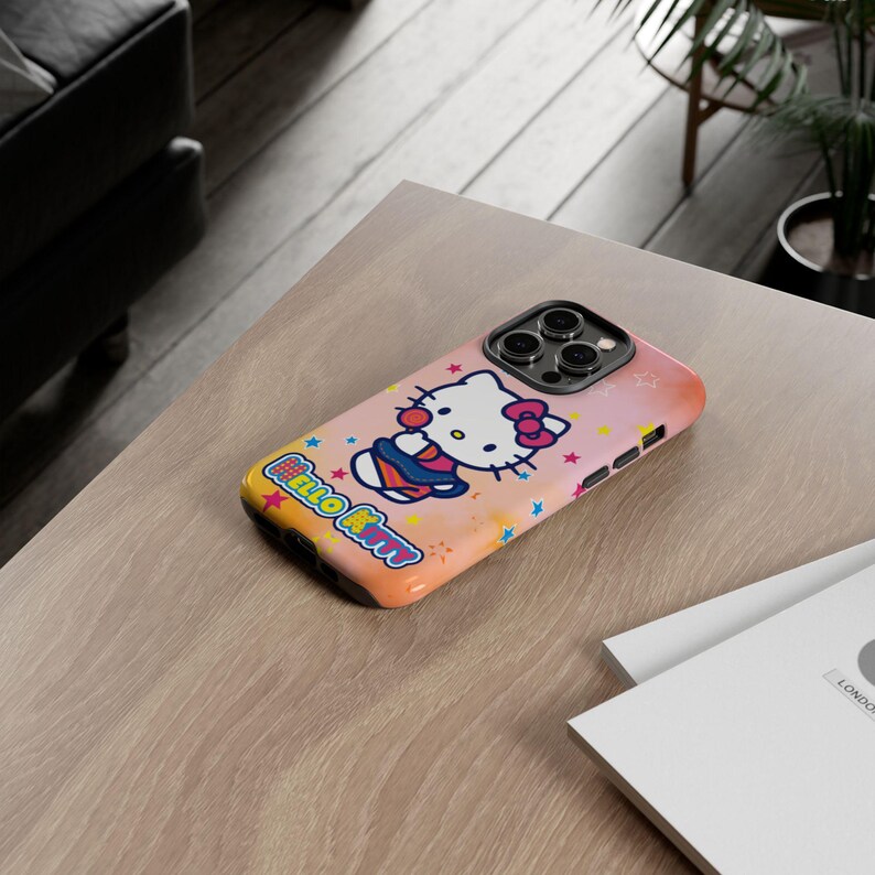 May include: A pink and orange iPhone case featuring Hello Kitty. The case has a colourful design with stars and the words "Hello Kitty". The phone case is on a light brown wooden table.
