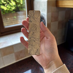Puede incluir: Un marcapáginas de madera con un diseño de mapa topográfico. El marcapáginas es rectangular con un pequeño agujero en la parte superior. El diseño del mapa presenta líneas negras curvas sobre un fondo marrón claro. El marcapáginas se sostiene en una mano.