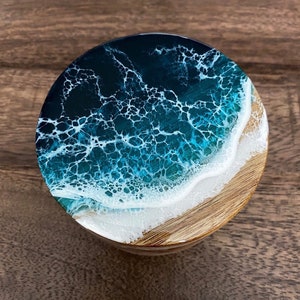 Könnte beinhalten: Ein runder Holz-Untersetzer mit einer Ozeanszene aus Harz. Das Harz ist klar und blau, mit weißen Schaumwellen am Rand. Der Untersetzer ist perfekt, um Ihrem Zuhause einen Hauch von Strand zu verleihen.