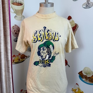 May include: Cream-colored t-shirt featuring the word "Genesis" in a blue and yellow font. The shirt displays a graphic of the Mad Hatter from Alice in Wonderland. The Mad Hatter is wearing a green hat and bow tie.