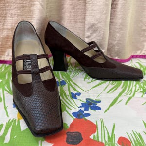 以下が含まれることがあります： 茶色の革とスエードのヴィンテージハイヒール。つま先にはヘビ柄、Tストラップのデザイン。ヒールは濃い茶色で、花柄の生地の上に置かれています。