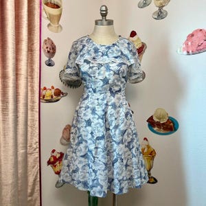 May include: A white dress with a blue floral pattern and a ruffled neckline. The dress has short sleeves and a flared skirt.