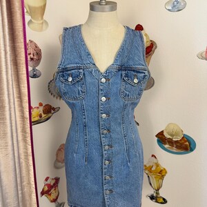 May include: A light blue denim dress with a V-neckline and button-down front. The dress features two chest pockets and a fitted silhouette. The dress is displayed on a mannequin. The background includes decorative ice cream images.