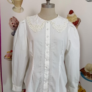 Op de afbeelding: Een witte blouse met een kanten Peter Pan kraag en lange mouwen. De voorkant heeft een knoopontwerp met verticale plooien en decoratieve kanten inzetstukken. De blouse wordt op een mannequin getoond.