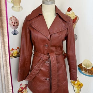 May include: A vintage, rust-coloured leather trench coat with a belted waist and button closure. The coat has a classic collar and long sleeves. The coat is displayed on a mannequin.