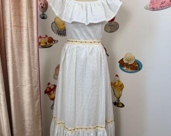 Puede incluir: Un vestido maxi blanco de encaje con cuello de volantes y un ribete amarillo y floral alrededor de la cintura y el dobladillo.