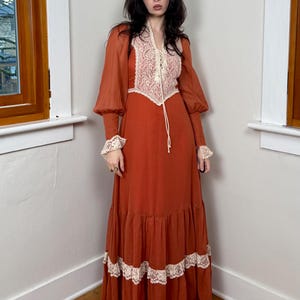 Könnte beinhalten: Ein rostfarbenes Maxikleid mit langen, durchsichtigen Ärmeln und einem mit Spitze besetzten Oberteil. Das Kleid hat einen Schnürhalsausschnitt und einen Stufenrock mit Spitzenakzenten. Die Ärmel haben gerüschte Manschetten und das Kleid hat einen Vintage-Bohemian-Stil.