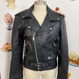 May include: Black leather biker jacket with a notched collar, an asymmetrical zip, and multiple zipped pockets. The jacket has a belt at the waist and silver-tone hardware. The jacket is displayed on a mannequin.