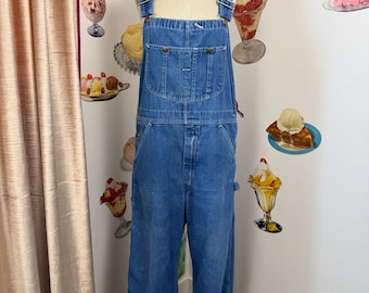 May include: Blue denim overalls with adjustable straps and multiple pockets. The overalls are displayed on a mannequin. The background features ice cream sundae decals and a pink curtain.
