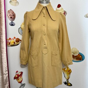 Pode incluir: Vestido vintage cor mostarda com gola grande, mangas compridas e dois bolsos frontais. O vestido tem uma frente abotoada e é exibido em um manequim. O vestido é na altura do joelho.