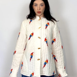May include: A cream-colored quilted jacket featuring a repeating pattern of colorful parrots in red, yellow, and blue. The jacket has a high collar, long sleeves, and is fastened down the front with four light brown toggle buttons.