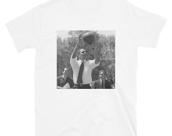 May include: A black and white photo of a man in a suit and tie, holding a basketball above his head. He is surrounded by trees and other people.