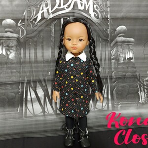 May include: A black and white doll with long black braids wearing a black dress with colorful polka dots and a white collar. The doll is standing in front of a black and white backdrop with a gate and the words "Addams" and "Kona Closet" visible.