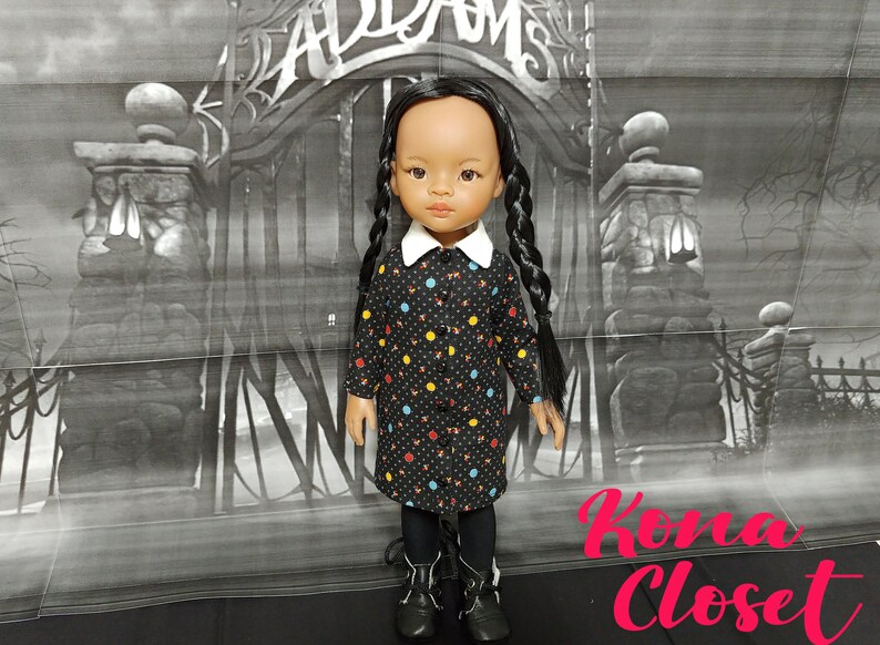 May include: A black and white doll with long black braids wearing a black dress with colorful polka dots and a white collar. The doll is standing in front of a black and white backdrop with a gate and the words "Addams" and "Kona Closet" visible.