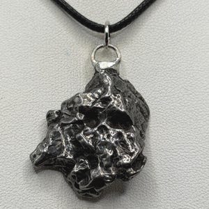 May include: A dark grey, metallic meteorite pendant with a rough, irregular, and pitted surface texture. The pendant is suspended from a silver-toned metal bail and attached to a simple black cord necklace, creating a raw and natural appearance.