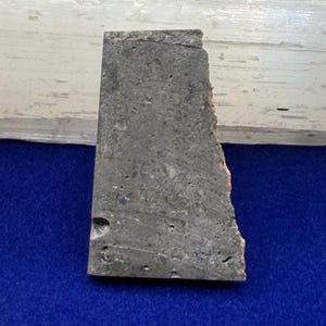 以下が含まれることがあります： 暗い灰色の石の台形型の破片で、ざらざらとした多孔質の質感と目に見える鉱物の含有物があります。この破片は、平らな上端、真っ直ぐな左側、そして基部近くに小さな円形のくぼみがあるギザギザで不均一な右端を特徴としています。