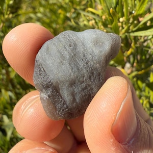 Op de afbeelding: Een grijze, onregelmatig gevormde edelsteen met een glad, gepolijst oppervlak. De steen heeft een licht iriserende glans.