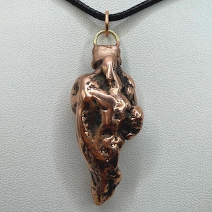 May include: A copper-colored pendant with an irregular, textured, and organic shape suspended from a thin black cord. The metal surface features a metallic sheen with dark, recessed areas and a polished, raised finish, attached by a gold-toned ring.