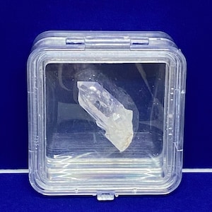 May include: A clear plastic box containing a clear crystal point. The crystal is a light purple colour and has a pointed tip.