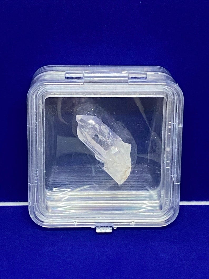 May include: A clear plastic box containing a clear crystal point. The crystal is a light purple colour and has a pointed tip.