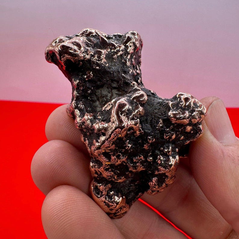May include: A rough, irregularly shaped, dark brown, copper nugget with a textured surface.