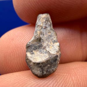 May include: A small, irregularly shaped stone fragment with a textured, weathered surface. The object displays a mottled grey and brown colour palette with visible pits, ridges, and uneven contours across its entire exterior, creating a rugged appearance.