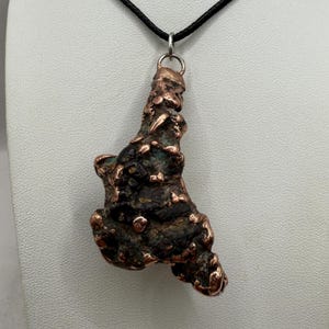 May include: A handcrafted copper pendant with a unique, textured, organic shape. The pendant is suspended from a black cord necklace. The copper has a dark brown and reddish-brown patina with hints of green.