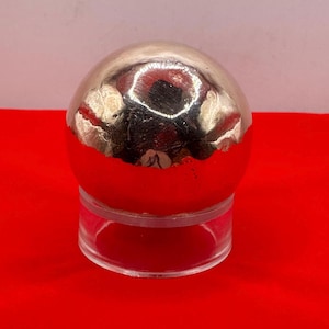 May include: A silver sphere on a clear acrylic stand. The sphere has a smooth, reflective surface with some imperfections.