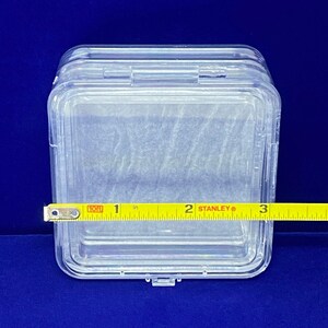 May include: A clear plastic storage box with a hinged lid. The box is approximately 5 cm wide.