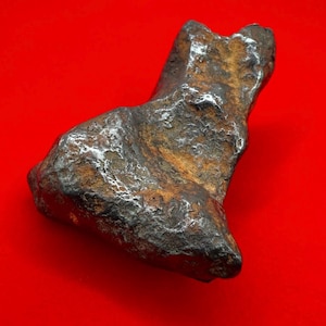 May include: A weathered, irregularly shaped, metallic object with a rough, textured surface. The object has a dark brown and silver colour scheme, with a reddish background. The object appears to be a rock or mineral specimen.