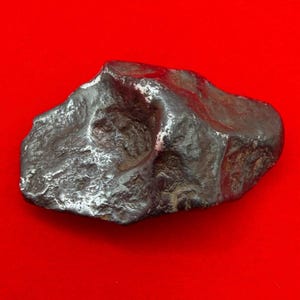 May include: A dark gray, metallic iron meteorite with a highly irregular, pitted surface texture. The object features deep indentations and smooth, rounded edges, showcasing a dense, heavy appearance with a reflective, gunmetal-colored finish.