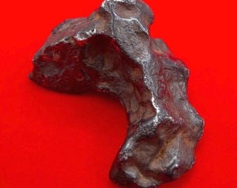 May include: A dark brown and metallic gray iron meteorite specimen with a highly irregular, pitted, and textured surface. The object features deep indentations and a rugged, organic shape with a dense, heavy appearance and a solid, weathered exterior.