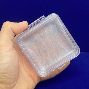 May include: A clear plastic square box with a hinged lid. The box is empty and has a white paper liner.
